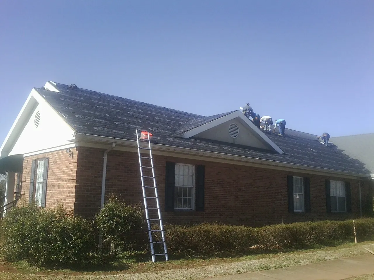 Crew tearing shingles off a brick ranch roof with a ladder set — Emergency Roof Repair work by our crew in Roma