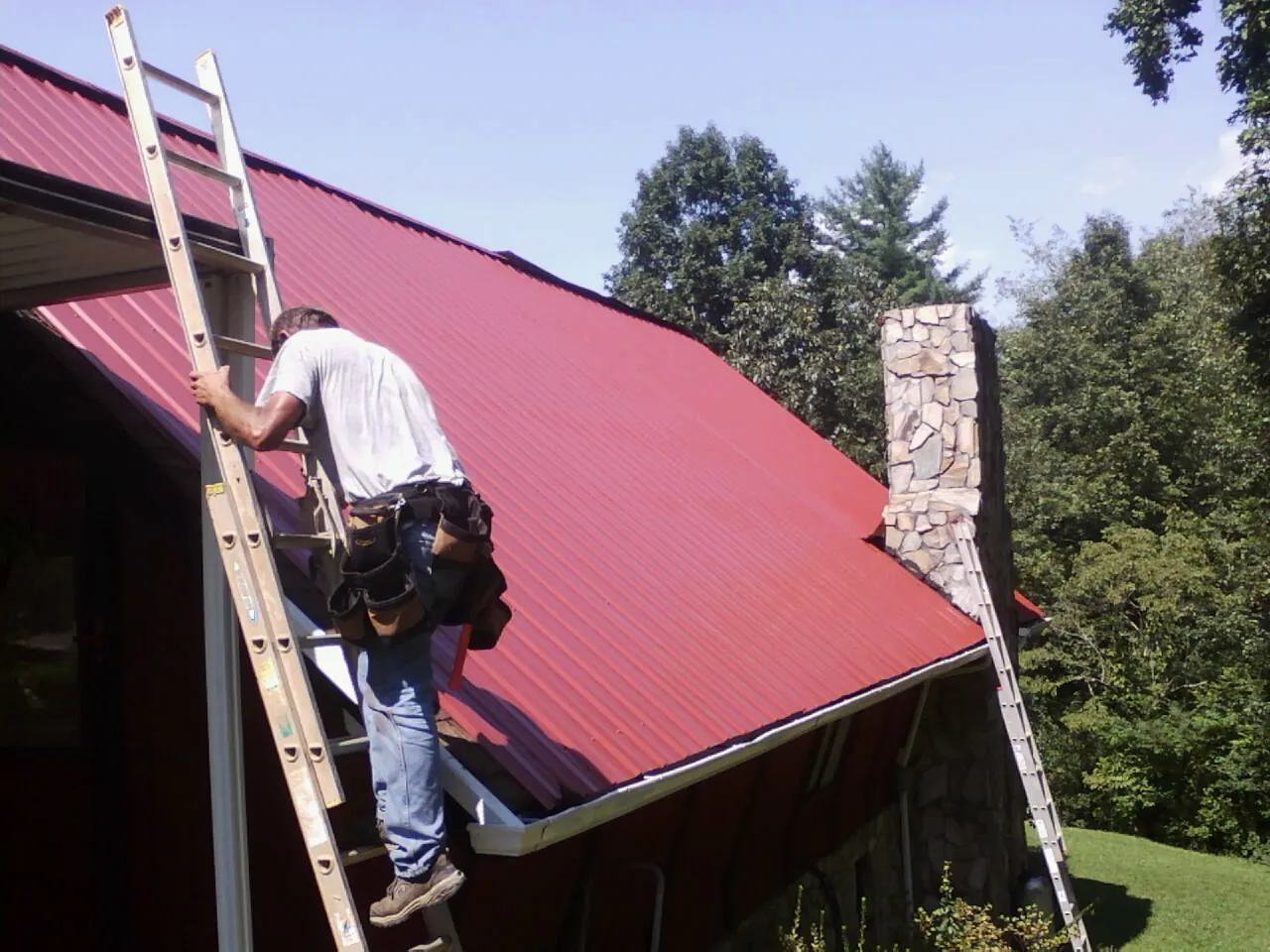 Roofer on a ladder beside a red metal roof and stone chimney — Soffit & Fascia Repair by Roofing Brothers in Roma